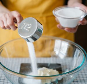 A cook using a half-cup dry measuring cup, pouring sugar into a large bowl.