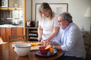 Couple is together at a dining table. Woman is slicing oranges on a cutting board.