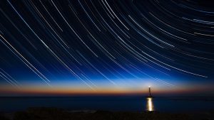 Stars, stretched out by long exposure, surround a lighthouse in a dark sea