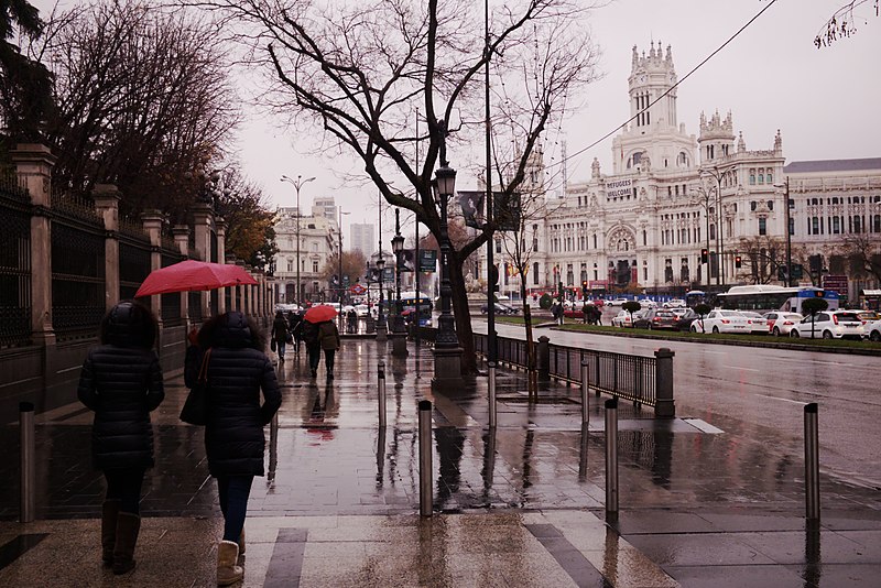 Several people with red umbrellas walk on a rainy sidewalk in Madrid, Spain. Across the street from them is a white cathedral.