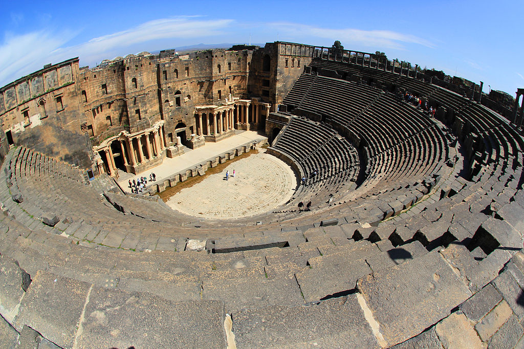 Panoramic view of Bosra amphitheater in Syria, showcasing ancient ruins against a blue sky backdrop.