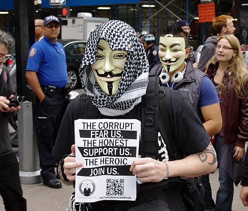A photo of a downtown area with passersby and two members of the group known by the pseudonym "Anonymous", at the Wall Street occupation protest in New York on September 17, 2011. Both members are wearing a "Guy Fawkes" type mask, and one is holding leaflets with both hands that read "The corrupt fear us, the honest support us, the heroic join us."