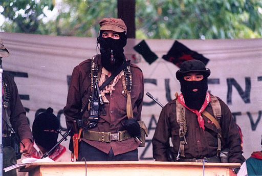 A photo showing two masked and armed men clothed in guerilla-type button-down shirts, bandanas and hats, speaking behind a lectern to an outdoor audience; Subcomandante Marcos and Comandante Tacho in La Realidad, Chiapas. The photo is somber, with an outstretched fabric sign in the background and other masked men.