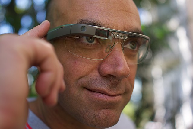 A white man wearing a pair of Google Glass smart glasses. The glasses have a sleek, modern design with a small rectangular display over the right eye. The background is blurred, focusing attention on the glasses and the person's face.