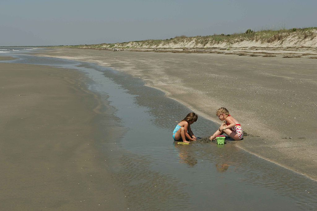A photo of two young children wearing swimsuits, playing in the sand near the water's edge on a deserted beach. One child, in a blue swimsuit, is digging with a yellow shovel, while the other, in a red polka-dot swimsuit, is using their hands to build something. The beach stretches out into the distance with grassy dunes visible in the background under a clear sky.