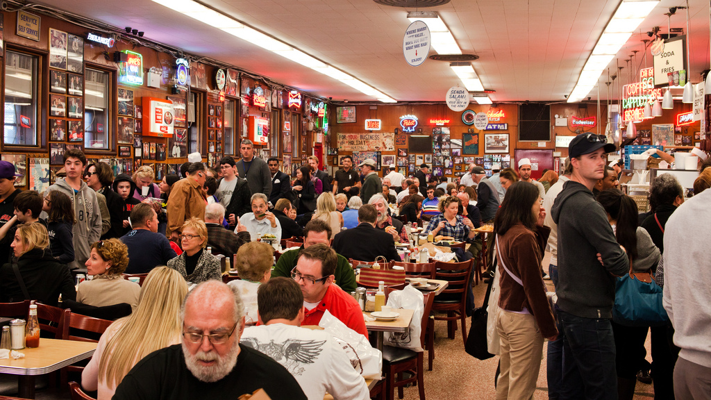 A crowded room with a couple dozen people at Katz's Deli in New York City with some people sitting at tables talking and eating, and others standing and waiting to receive or place their orders. The walls are filled with framed pictures and some neon signs.