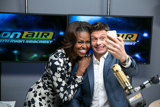 First Lady Michelle Obama poses for a selfie with Ryan Seacrest in his radio studio. There are two tv screens behind them, the walls are grey, and a microphone is suspended in front of them.