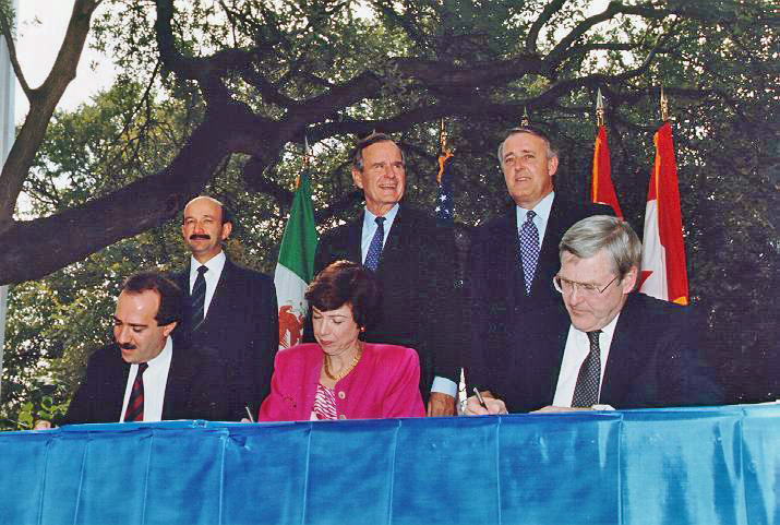 A photo two men and one woman, seated at a table with blue cloth, each signing documents. Behind them stand three prominent leaders of Mexico, the U.S., and Canada during NAFTA treaty signing in San Antonio, Texas, Oct. 7, 1992. From left to right: Mexican president Carlos Salinas, US president George H.W. Bush and Canadian prime minister Brian Mulroney. They are positioned in front of their respective national flags. The setting is outdoors, with large tree branches visible in the background, suggesting a formal ceremony in a park or garden. The atmosphere is serious and ceremonial, highlighting the importance of the occasion.