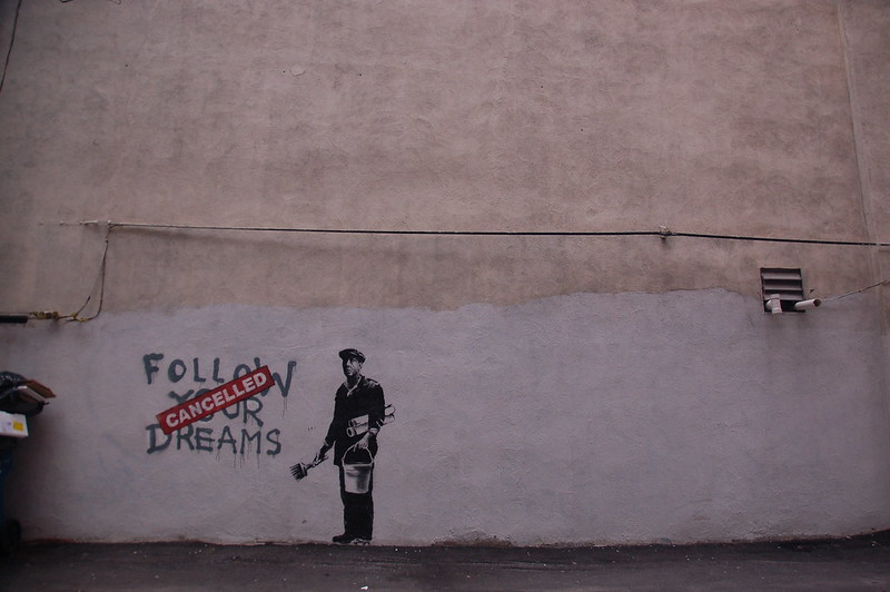 Street art by Banksy located in the Chinatown area of Boston, depicting a man holding a paint brush and bucket standing by a spray-painted sign saying 'follow your dreams'. The drip-dried slogan, which is dark grey along with the man, had a red stamp over it reading in all capital letters, 'cancelled'.