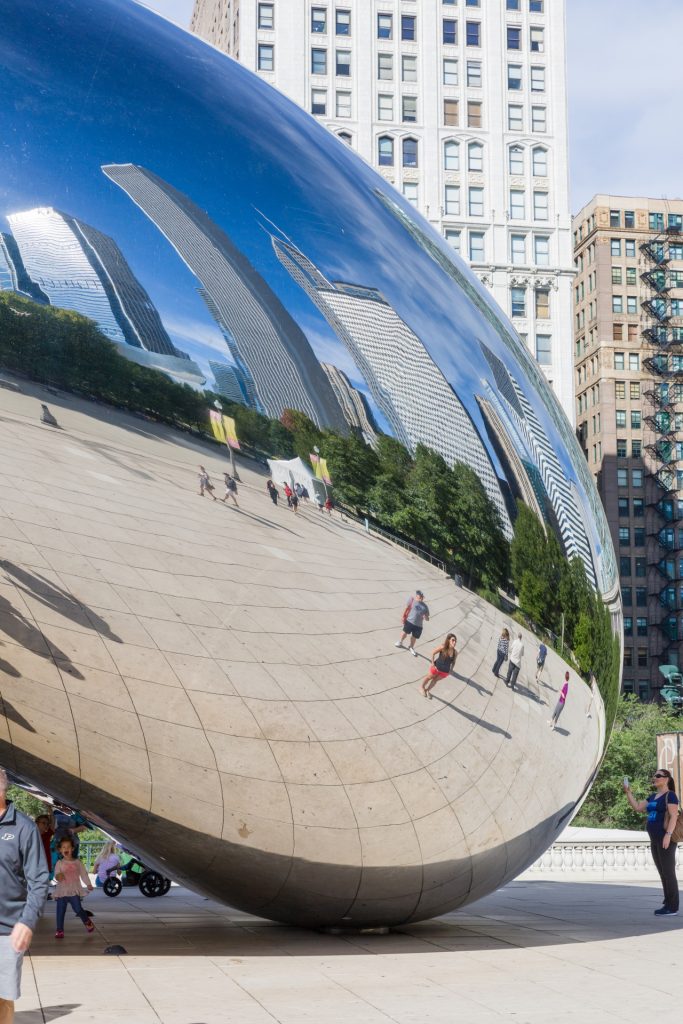 Photograph of a large, reflective sculpture known as "Cloud Gate" or "The Bean" in Chicago's Millennium Park. The sculpture's polished surface mirrors the surrounding skyscrapers, trees, and people walking nearby. The reflections are slightly distorted, creating a funhouse mirror effect. Visitors are seen strolling around the sculpture, taking photos, and enjoying the sunny day. Tall buildings frame the background under a clear blue sky.