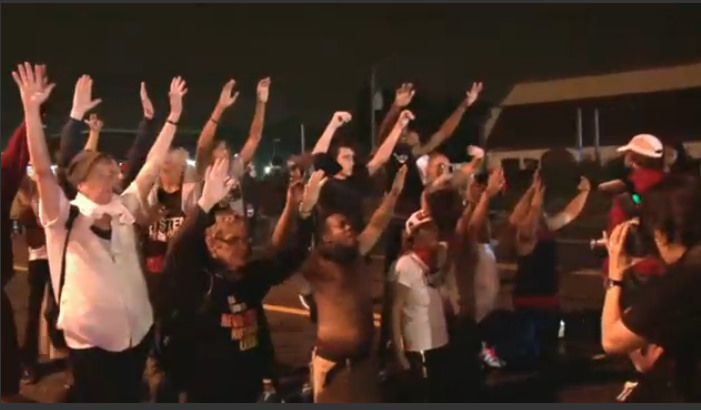 A nighttime scene of protestors standing a kneeling with their hands up while photographers in front of them snap photos. They are protesting police violence with this gesture of "hands up, don't shoot."