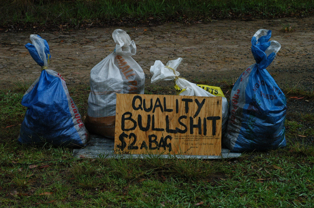 Outdoor image of four, tied bags of trash (two white, two blue) o grass with a plywood sign in front of them reading "quality bullshit $2 a bag".