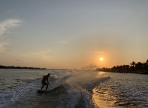Picture of a man wakeboarding behind a boat with the setting sun reflecting off the wave and palm trees in the background.