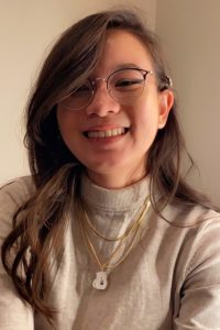 A photo of contributing author, Jacquie Kuru. She is smiling, wearing wire-framed glasses, gold necklaces, a beige turtleneck top, and long, brown hair with soft waves and bangs.
