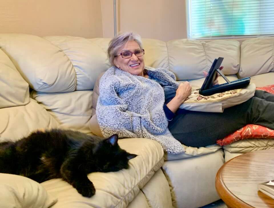 Picture of a grey-haired woman with reading glasses, wrapped in a heather grey, faux-shearling jacket, reclining on a cream-colored leather sectional couch in a living room, with a black cat beside her, and holding an electronic device.